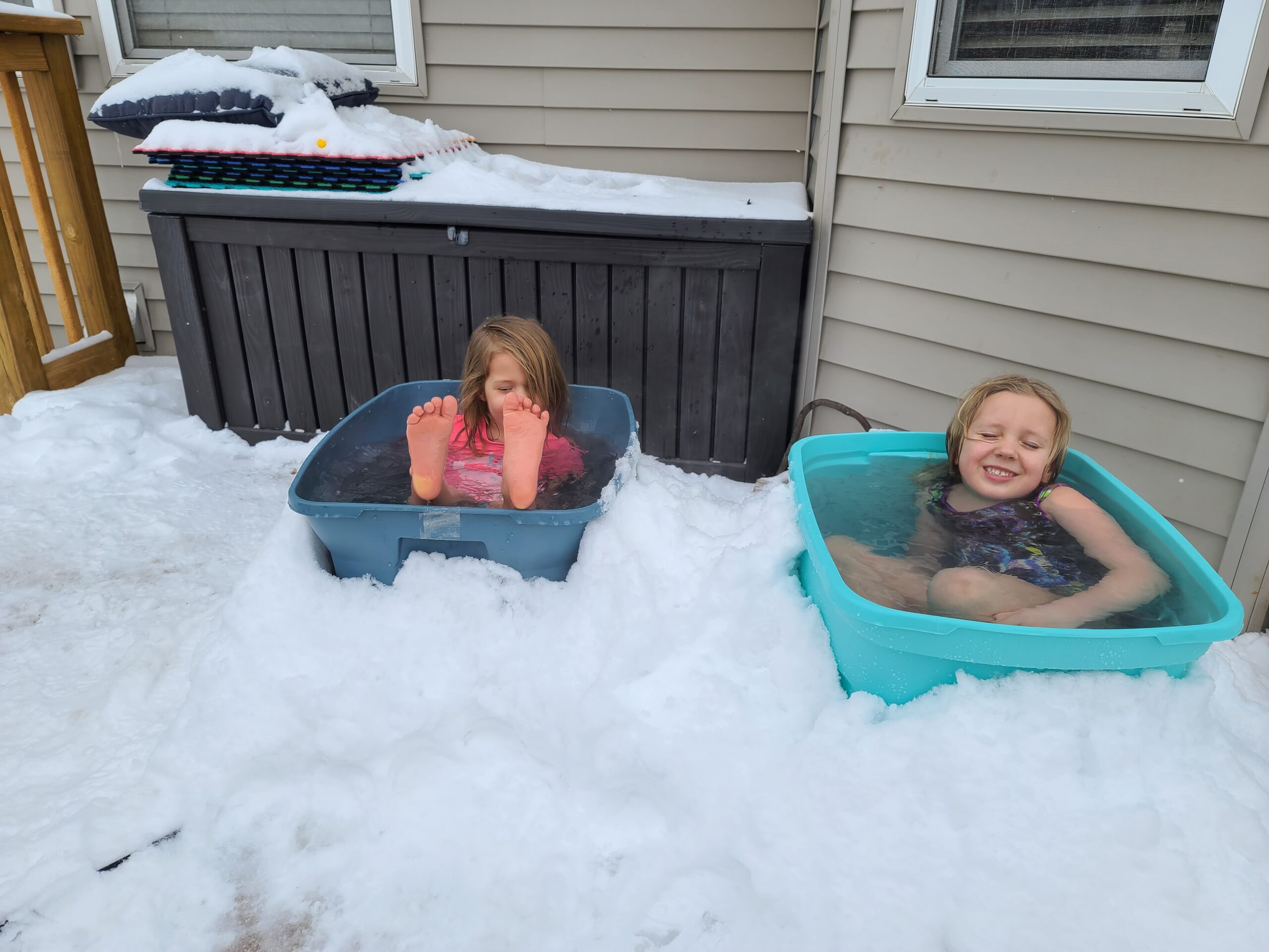 Two children are sitting in storage bins full of water. They're wearing bathing suits and smiling. The bins are surrounded by snow.